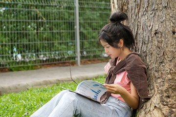 A high school student is sitting against a tree, deeply engrossed in her studies. She is focused on...