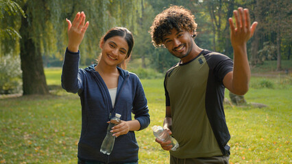 Two sport people smiling talking holding water bottles smile invitation join to exercise male Indian female Arabian friends couple man woman in park invite welcome workout fitness fit in city motivate
