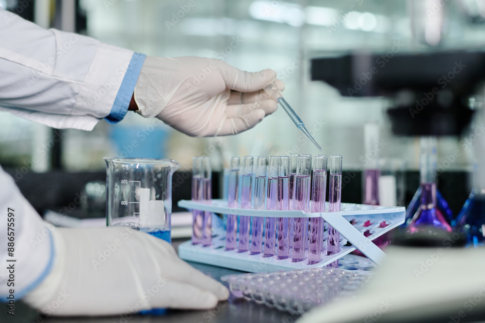 Poster Gloved hand of modern clinician standing by workplace in front of camera and dropping liquid substance into flasks from plastic pipette