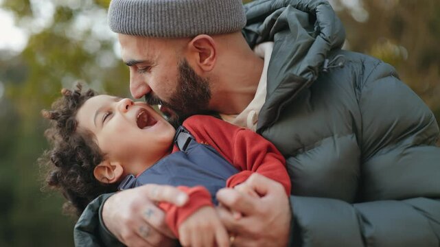 Close-up of a loving, bearded father sharing a kiss and playful moment with his delighted little son on a chilly day outside, surrounded by nature.