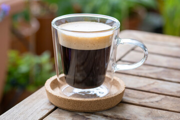 Cup of coffee with the foam in transparent glass on the wooden table in the garden.