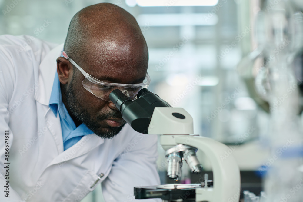 Poster Young serious scientist in protective eyeglasses looking in microscope while studying new strain or toxic chemical substance