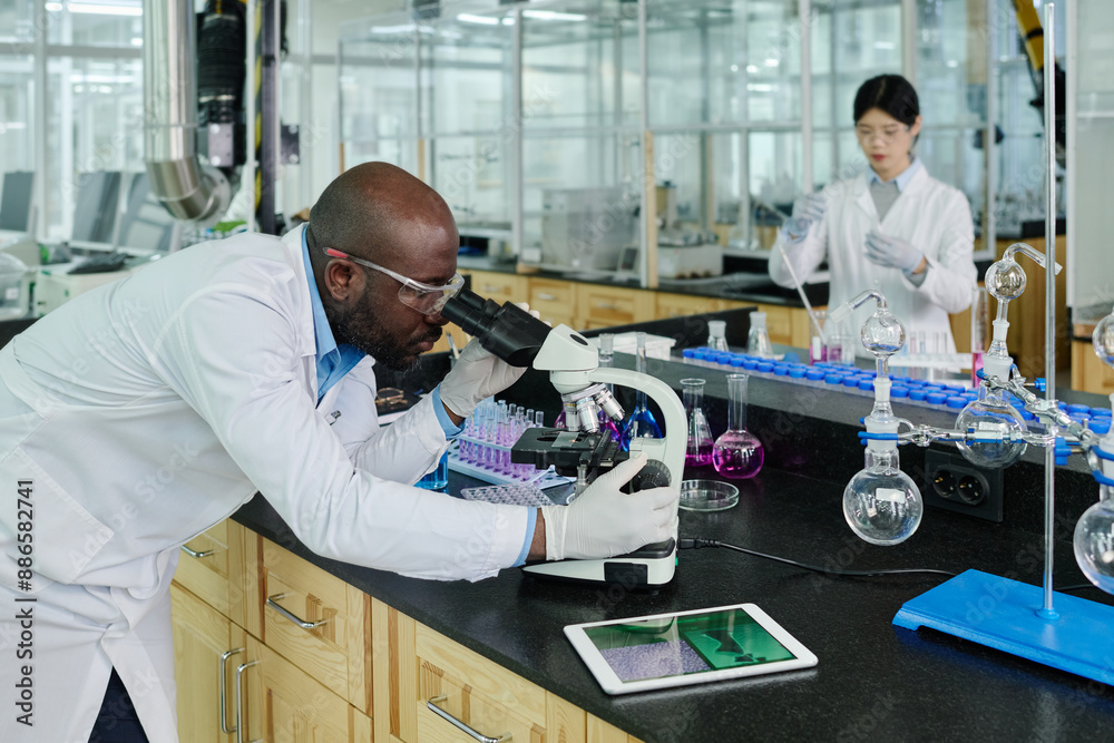 Aufkleber Side view of young scientist or virologist bending over microscope and researching new virus while working by table with glassware