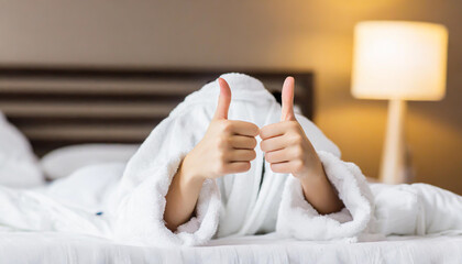 gesture, comfort and morning concept - hands of young woman in hotel robe lying in bed and showing thumbs up at bedroom