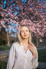 Fashion outdoor photo of beautiful woman with blond hair in elegant suit posing in spring flowering park with blooming cherry tree. Copy space and empty place for advertising text