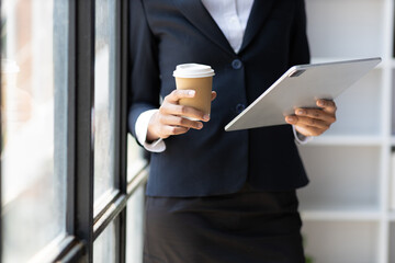 Businesswoman stands holding a tablet in the office and drinks hot coffee.