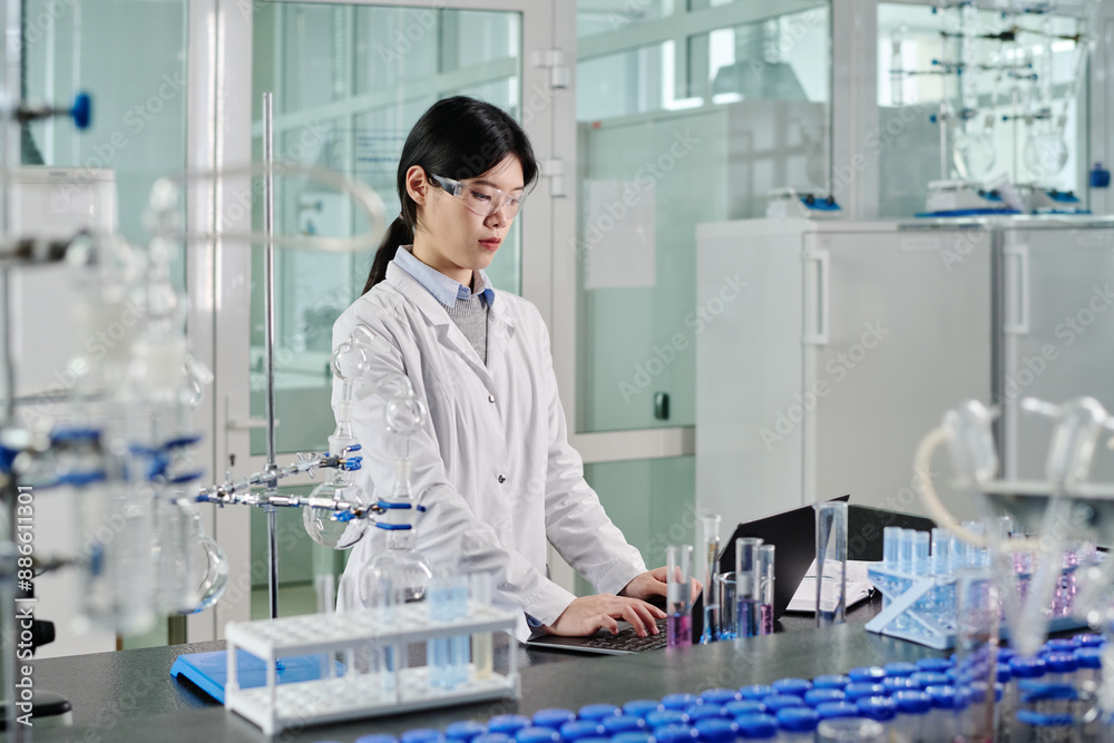 Aufkleber young asian female scientist in lab coat and protective eyeglasses looking at laptop screen and typi
