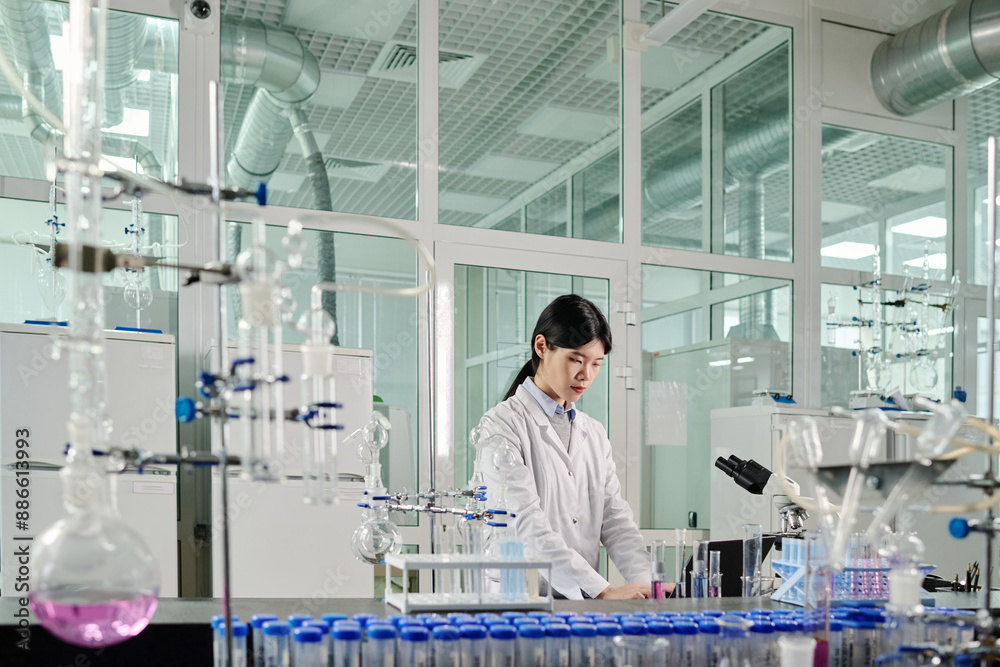 Poster young brunette woman in lab coat standing by workplace with glassware and other equipment and enteri