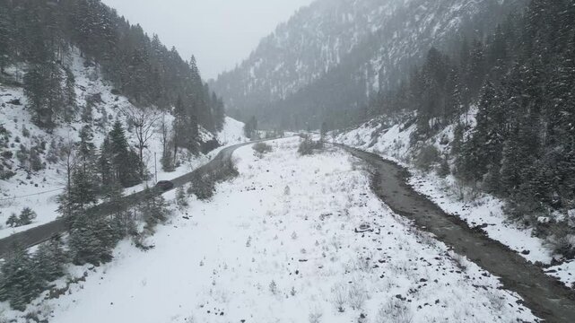 winter mountain valley cars driving forward aerial snow covered flocks falling white forest trees street road stream water