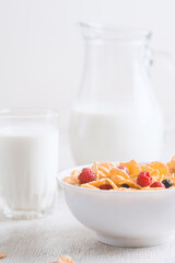 bowl of corn granola with milk, fresh raspberries, blueberries . milk in a jug and a glass  on white wooden board for healthy breakfast
