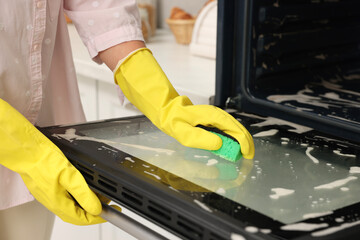 Woman cleaning oven door with sponge in kitchen, closeup