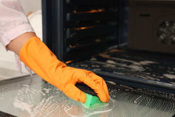 Woman cleaning oven door with sponge in kitchen, closeup