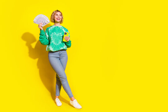 Full length photo of cheerful pretty lady dressed green sweatshirt winning money modern gadget empty space isolated yellow color background