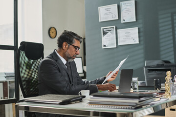 Side view of serious mature man in formal suit looking through juridical paper document and typing on laptop keyboard by workplace