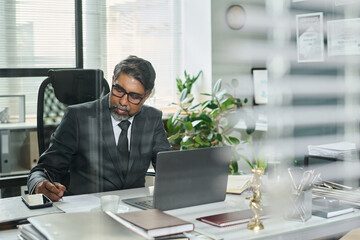 Obraz premium Serious mature male lawyer sitting by workplace in front of laptop, reading paper document and putting his signature below text