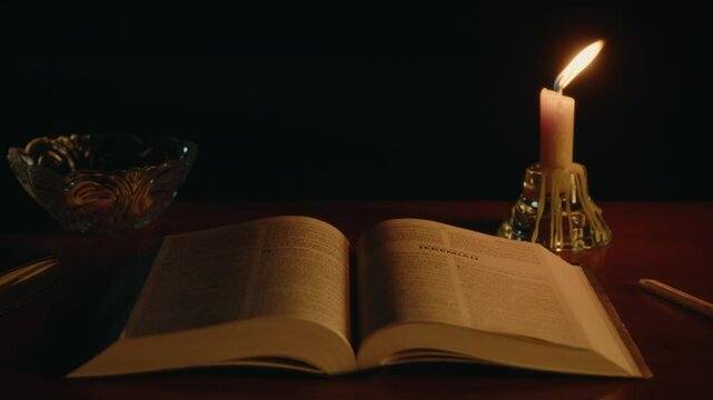Opened book on a desk, Bible in soft candle light, candle is burning nearby, spiritual practice, High quality 4k footage