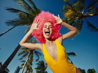 A woman with pink afro hair in a yellow swimsuit stands in front of multiple palm trees, creating a...