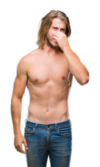 Fototapeta premium Young handsome shirtless man with long hair showing sexy body over isolated background smelling something stinky and disgusting, intolerable smell, holding breath with fingers on nose.