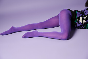 A young woman with vibrant tights and sweater poses gracefully on a purple background in a studio setting.