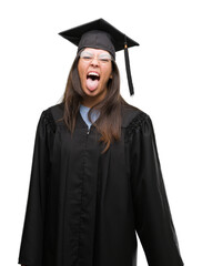 Young hispanic woman wearing graduated cap and uniform sticking tongue out happy with funny expression. Emotion concept.