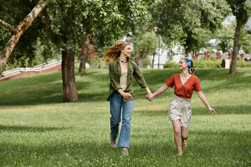 Fototapeta premium Two young women holding hands and walking through a lush green park, smiling and laughing together.