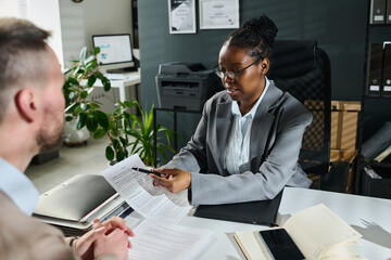 Confident African American businesswoman or lawyer pointing at title of paper document while explaining male client its main points