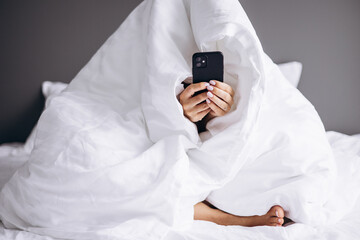 Woman sitting in bed covered with blanket and using mobile phone