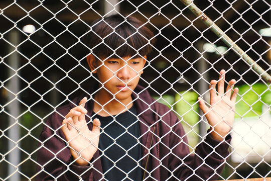 Asian male teenager standing on iron fence in juvenile detention center. Concept of detention, freedom, imprisonment, confinement, reformation, loneliness.