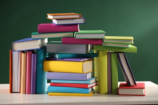 Stacks of many colorful books on white wooden table near chalkboard in classroom