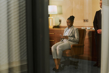 Young African American female financial director or chief executive officer in formal suit sitting in armchair in boardroom and texting