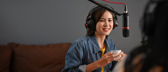 A woman actively engaging with her guest during a podcast recording, holding a cup and using professional microphones, set in a relaxed studio environment with copy space