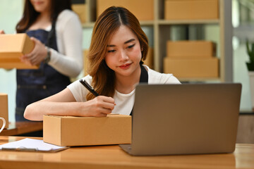 A woman uses a laptop to manage online orders while writing an address on a package, with another person in the background handling more boxes, integrating digital and manual processes in SME