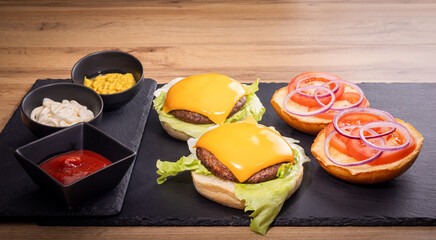 Gourmet burgers on a black slate platter featuring juicy beef patties topped with melted cheddar cheese, fresh lettuce, and sliced tomatoes and red onions, with bowls of ketchup, mayo and mustard.