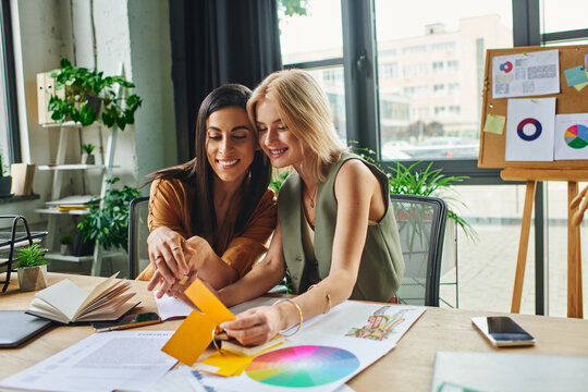 Two women, lesbian couple, collaborate on a project in a modern agency workspace, discussing color palettes and designs.