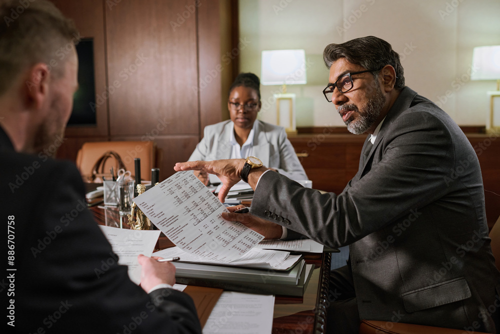 Wall mural Confident aged male director of lawyer company looking at one of two intercultural subordinates during discussion of paper document