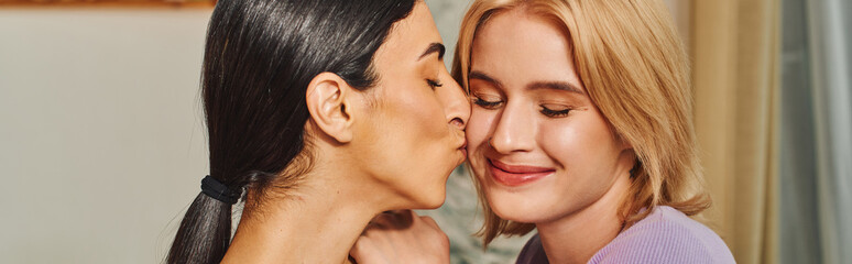 Two women, one with dark hair and the other with blonde hair, hug and kiss in a cozy home setting.