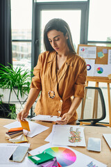 A woman in a mustard shirt examines a design sketch at a cluttered desk with color palettes,...