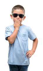 Dark haired little child wearing sunglasses cover mouth with hand shocked with shame for mistake, expression of fear, scared in silence, secret concept