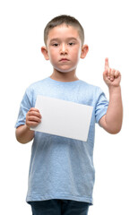 Dark haired little child holding a blank card surprised with an idea or question pointing finger with happy face, number one