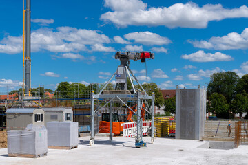 A construction crane is lifting a large concrete block at a building site. The crane is mounted on a platform, and there are other construction materials and equipment visible in the background.