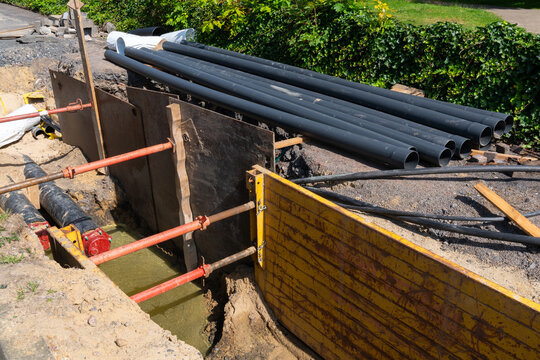 This photo depicts a trench at a construction site, secured with shoring to prevent collapse. Several large pipes are stacked nearby, ready for installation. The trench is partially filled with water