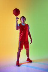Confident and ready, young basketball player in red stands with ball in neon light against gradient...