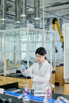 Chinese female researcher in lab coat, protective eyeglasses and gloves carrying out chemical investigation while standing by workplace
