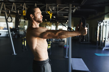 A muscular man without a shirt is holding a barbell in a gym, showcasing his strength and...