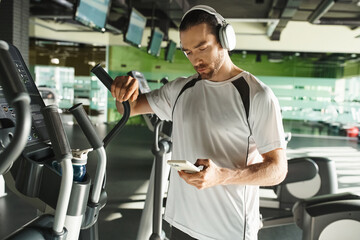 A focused man in athletic wear wearing headphones and using a cell phone while working out in a gym.