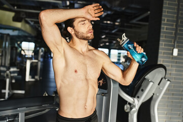 Naklejka premium Strong shirtless man taking a break, holding a bottle of water in a gym setting.