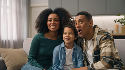 Excited happy playful African American ethnic family mother father embracing little boy child playing game parents kid watching funny TV show movie posing making faces grimacing indoors home apartment