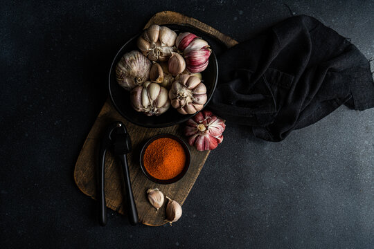 Overhead view of a bowl of fresh garlic bulbs on a chopping board with garlic cloves, chilli pepper salt and a garlic press