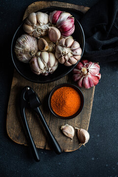 Overhead view of a bowl of fresh garlic bulbs on a chopping board with garlic cloves, chilli pepper salt and a garlic press