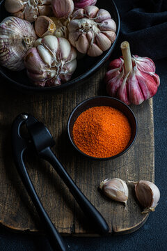 Overhead view of a bowl of fresh garlic bulbs on a chopping board with garlic cloves, chilli pepper salt and a garlic press